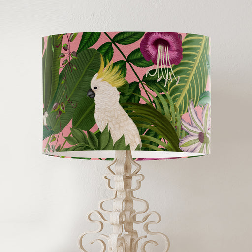 Parrots in white, green & blue along with monkeys sit amoungst green tropical leaves on a pink background on a classic sized 30x21cm handcrafted fabric lampshade by artist Kelly Stevens-McLaughlan